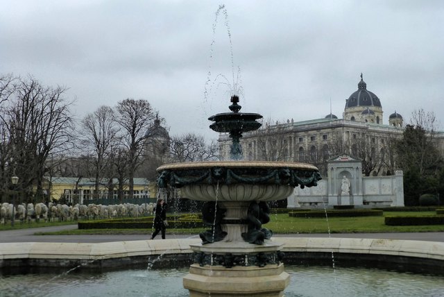 Im Volksgarten. Die Rosenstöcke haben immer noch ihre winterlichen Mönchskutten an, aber der Volksgarten-Brunnen darf schon plätschern. Im Hintergrund das Grillparzer-Denkmal und die beiden großen Museen.