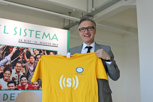 Vorstandsvoritzender Werner Binnenstein-Bachstein zeigt stolz das superar-T-Shirt. Auf der Rückseite steht das Programm: "sing.dance.succeed"