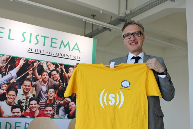 Vorstandsvoritzender Werner Binnenstein-Bachstein zeigt stolz das superar-T-Shirt. Auf der Rückseite steht das Programm: "sing.dance.succeed"