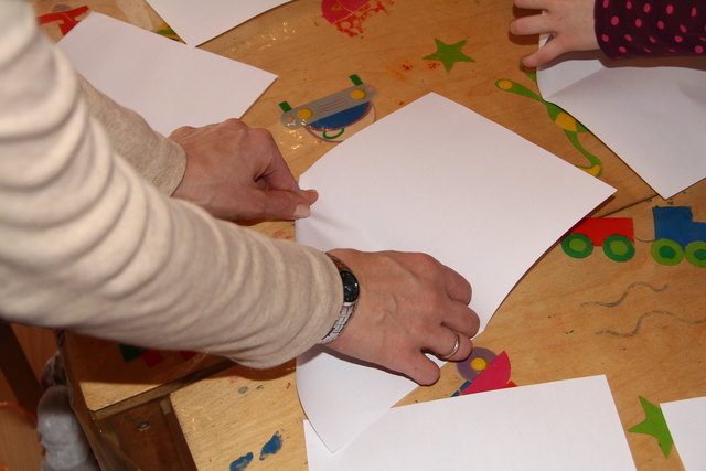 Beim Falten wirst du möglicherweise etwas Hilfe brauchen. Vielleicht können dir deine Eltern etwas zur Hand gehen. Lege ein DIN A4 Blatt im Hochformat vor dir auf den Tisch. Nimm die untere Seite und falte sie zur oberen. So entsteht in der Blattmitte ein Bug. Dann wird die erste Ecke  wie auf dem Bild entlang des Bugs eingefaltet.