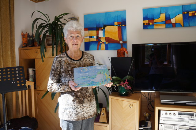 Elfriede  Benesch mit ihrem Kinderbuch, im Hintergrund zwei ihrer selbst gemalten Bilder.