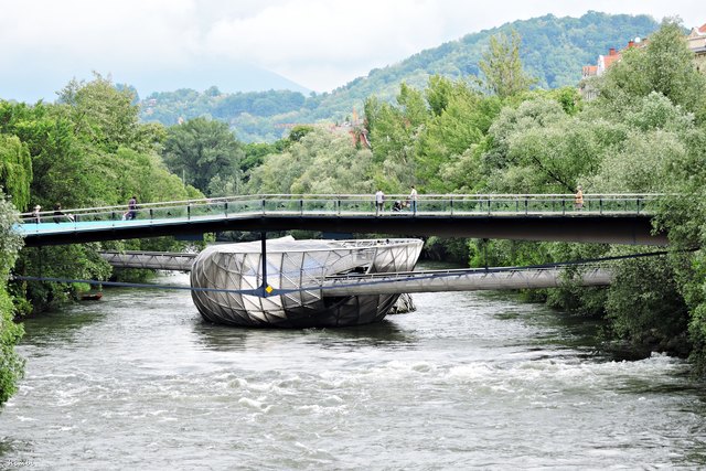 Die Murinsel Graz wurde anlässlich des Kulturhauptstadtjahres 2003 in Auftrag gegeben. Was sich Insel nennt, ist korrekterweise ein Schiff. Es hängt an einem Anker und wird durch die Stege zu den beiden Ufern zusätzlich stabilisiert.