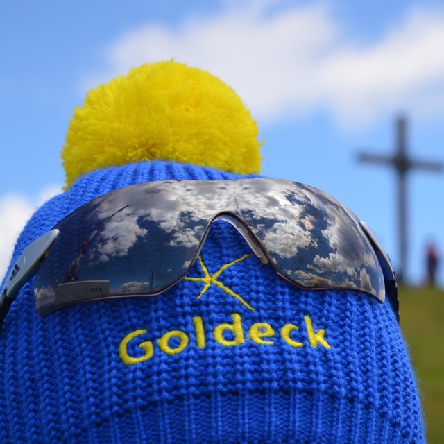 Goldeck-Pudelhaube mit Wolkenmuster auf der Sonnenbrille und Gipfelkreuz im Hintergrund.