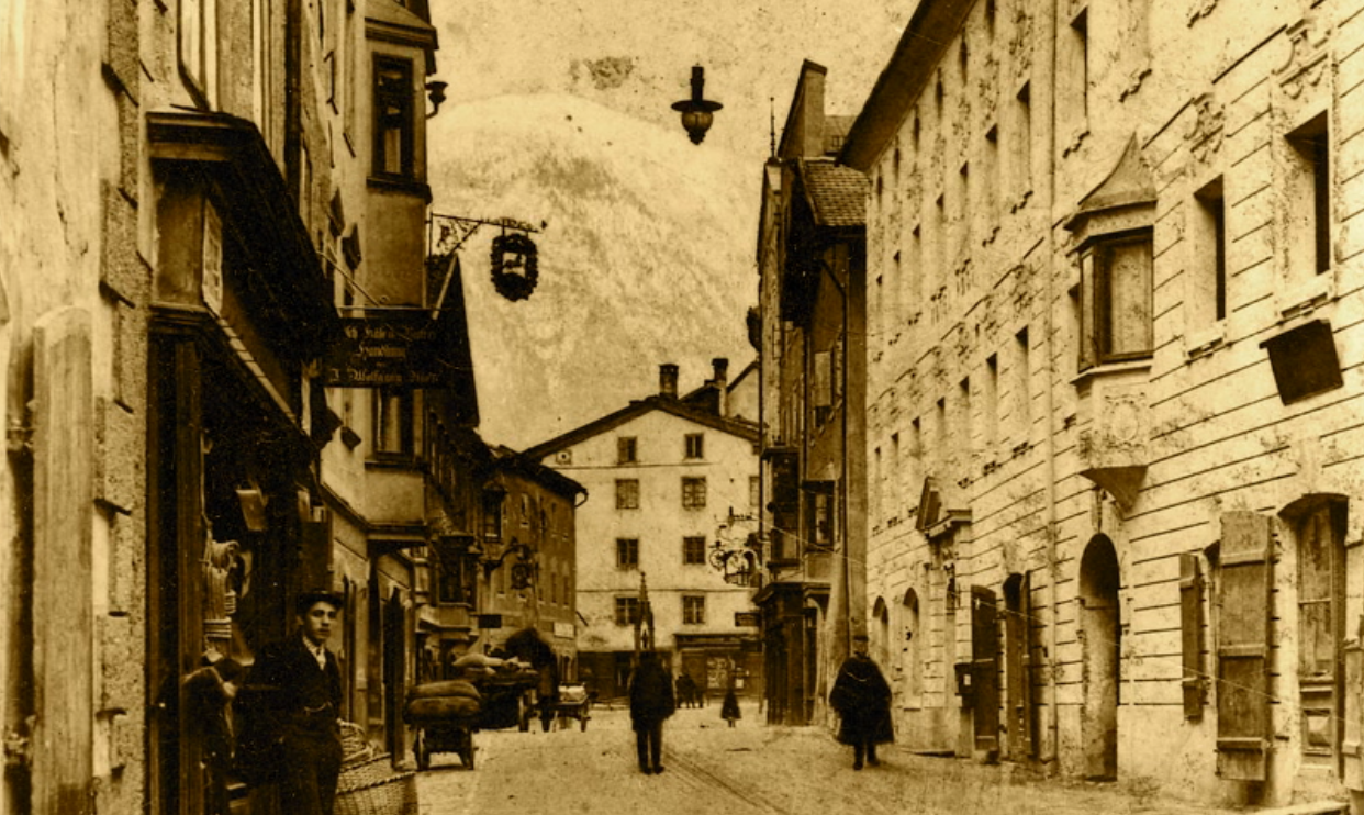 Die einstige Sigismundstraße (heute Innsbrucker Straße) auf Höhe ehem. Gasthof „alte Post“ (heute Raiba) und GH Weißes Rössl (GH Lorenzetti, heute Kögl) mit der 4. Bogenlampe damals im Markt Schwaz mit Blick zum Stadtplatz und einer Wasser-Rinne mitten auf der Schotterstraße noch ohne Gehsteige.