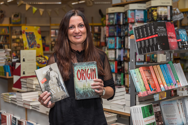 Susanne Rothberger von Frick greift zu empfehlenswerten Büchern und weist auf die Fülle der aktuellen Neuerscheinungen hin, die auf einem großen Tisch und Regal zur Auswahl stehen.