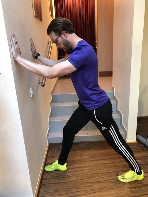 Mit aufrechtem Körper mit den Händen an der Wand abstützen. Die Fersen sind am Boden und das Bein an der Wand ist leicht abgewinkelt.