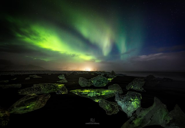 Smaragde am Diamanten Strand 

Gleich neben der berühmten Jökulsarlon-Gletscherlagune findet sich der schwarze Vulkansandstrand, wo die Eisberge aus der Lagune an Land treiben und einen magischen Ort erschaffen. Die blauen, schwarzen, weißen, durchsichtigen und manchmal auch grünen Eisberge liegen am Strand und ordnen sich bei jeder erneuten Flut neu an.

Als wir Mitten in der Nacht zum Strand gekommen sind, schwemmte die Flut wieder neue Eisschollen an. Wir waren ja schon am Vortag hier, mit einem komplett anderen Vordergrund. Die Suche nach einem Vordergrund dauerte nicht lange und wir fingen mit dem Fotografieren an. Die Wolkendecke öffnete ein etwas größeres Fenster und auch die Nordlichtaktivität war günstig. 
Ich machte das erste Foto zum Einstellen der Komposition, ein zweites Foto und plötzlich kam wie aus dem Nichts eine große Welle. Ich stand eher am Ende des Strandes und dachte nicht das es so weit Außen, so große Wellen herschaffen - falsch gedacht. Die Welle füllte jeden unserer Stiefel mit Wasser, da konnten wir noch so schnell laufen. Ich kann mich noch genau Erinnern als wir schreiend, jeder mit seinem Stativ in der Hand Richtung Festland flüchteten. 
Nachdem wir unsere Stiefel entleert haben, war es dann auch vorbei mit den Nordlichtern - zumindest für diese Nacht. 

Nordlichter waren ziemlich rar bei unserer Reise in Island. Zumindest hatte ich diese Nacht Glück mit diesem Foto.
Die Eisschollen haben die Farbe der Nordlichter wunderschön reflektiert und erinnern mich an Smaragde. Ein Fokus Stacking ging sich aber leider nicht mehr aus - bedingt durch die Welle. Ich hoffe das Bild gefällt euch trotzdem.