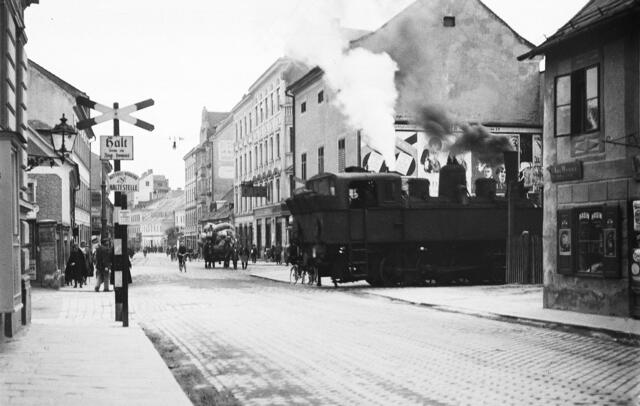 Damals 1930: Eine Dampflok fährt durch Urfahr
Im Jahr 1930 fuhr noch eine Dampflok durch Urfahr, wie hier an der Kreuzung Hauptstraße – Reindlstraße. Nach der Eröffnung der Mühlkreisbahn, die heute mit Dieselantrieb fährt, im Jahr 1888 ging die Verlängerung zum damaligen Staatsbahnhof Linz in Betrieb. Voraussetzung dafür war die seit 1900 bestehende Eisenbahnbrücke. Die Hauptstraße zeigte sich als „autofreie Zone“ mit Pferdefuhrwerken und Radfahrern. | Foto: Archiv der Stadt Linz