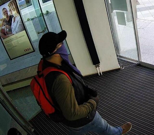 Der gesuchte Bankräuber: maskiert mit auffälligen Halstuch in länglicher Form (violett/bläulich mit rundem Logo, Schriftzug und 2 Bändern zur rückseitigen Befestigung im Kopfbereich), Sonnenbrille und schwarzer Baseballkappe. Der Täter trug einen roten Rucksack.