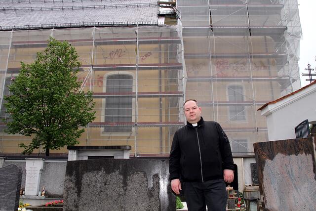 Pfarrer Markus Lehr vor der zurzeit bis zur Turmuhr eingerüsteten Pfarrkirche. Auf der Außenwand hat sich ein ungebetener „Künstler“ ausgetobt...  | Foto: Josef Fürbass