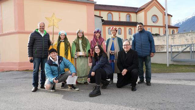 Der Telfer Pfarrer Peter Scheiring (links stehend), die Sternsinger Suara, Jonas, Mona und Chantrea (v.l.n.r.), Vikar Tony Bharath Kenneth Matthew (rechts stehend), die Sternsingerbegleiter Paula Lorenz und Felix Frick (vorne kniend), Bischof Hermann Glettler (vorne kniend) und die heimische Basilika im Hintergrund. | Foto: Cincelli/dibk.at