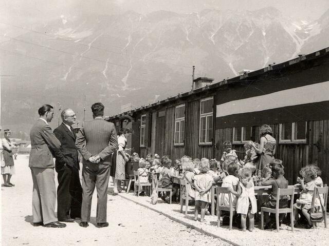 Kindergarten Reichenau. Bürgermeister Dr. Anton Melzer und Begleiter besuchen den Baracken-Kindergarten Reichenauer Lager, die Kinder sind gerade beim Jausnen, im Hintergrund ist die Nordkette. 1946. | Foto: Stadtarchiv/Stadtmuseum