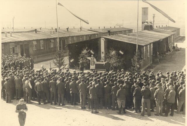Lager Reichenau mit Kriegsgefangenen nach dem Zweiten Weltkrieg. Feldkaplan Josef Klotz liest gerade vor den Holzbaracken eine Hl. Messe. Auf den Fahnenstangen die Tiroler Fahne, auf der Barackenwand die österreichische Fahne. Der Altarraum mit Nadelbäumen "geschmückt". 1945-1947. | Foto: Stadtarchiv/Stadtmuseum