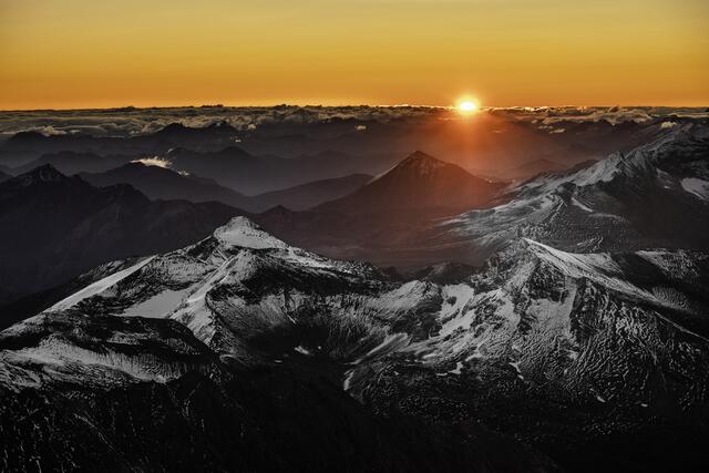 Der Zauber des Sonnenaufgangs am Gipfel des Großglockners. Der schneebedeckte Berg rechts ist die Racherin, links der Spielmann, dahinter das dunkle Dreieck des Brennkogels.