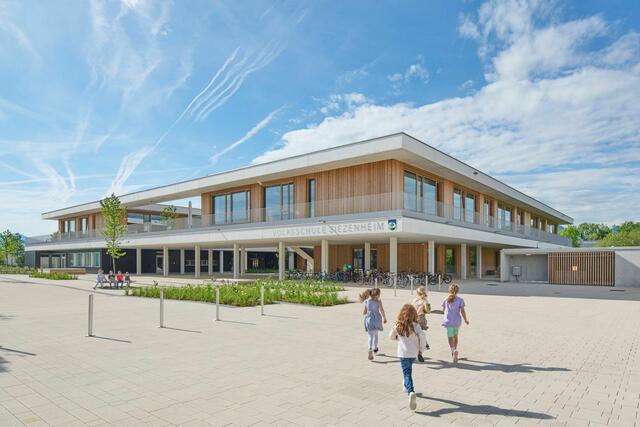 Siezenheim: Das neue Schulhaus, Resultat eines EU-weiten Architektur-Wettbewerbs, umfasst nicht nur eine Volksschule, eine Musikschule und eine auch von Externen nutzbare Turnhalle, sondern auch einen gedeckten Marktplatz und Jugendtreffpunkt im öffentlichen Raum. Hinter dem komplexen Raumsystem aus Clustern und Schulklassen steckt ein Hybridbau aus Stahlbetonskelett – teils in Recyclingbeton ausgeführt – und ausfachenden Massivholzwänden. Mit wenigen Handgriffen lassen sich die Klassenwände zur Seite schieben und ermöglichen auf diese Weise ein offenes Lernen. Außergewöhnlich ist auch die Energieversorgung: Obwohl die Biomasse-Nahwärme vor der Tür lag, entschied sich die Gemeinde aus ökologischen wie auch langfristig ökonomischen Gründen, auf Geothermie zu setzen. | Foto: Lukas Schaller