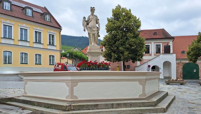 Der Brunnen in der Mitte des Kirchenplatzes wurde im dritten Viertel des 18. Jahrhunderts errichtet. Er hat ein achteckiges Becken und eine Brunnenfigur mit Palmblättern auf einem geschwungenen Sockel.