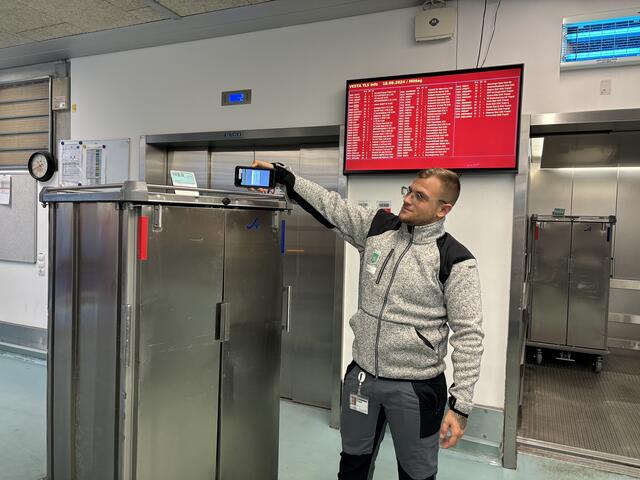 Logistikmitarbeiter Kevin Frick scannt den Barcode am Speisentransportwagen und sorgt somit dafür, dass die Patientinnen und Patienten auf der Urologie rechtzeitig ihr Mittagessen bekommen. Im Hintergrund ersichtlich ist der Screen, auf dem alle Transporte aufscheinen. | Foto: MeinBezirk