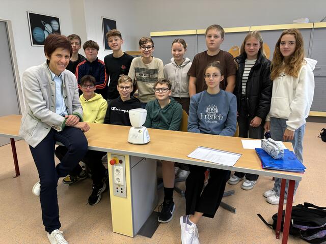 Schulleiterin Brigitte Reisinger (vorne links) mit einem Teil der 4b-Klasse des Gymnasiums Schärding und dem Avatar (am Tisch). Mit dessen Hilfe nimmt der an Long Covid erkrankte Schüler, wenn es ihm gesundheitlich möglich ist, am Unterricht teil. | Foto: MeinBezirk/Bichler