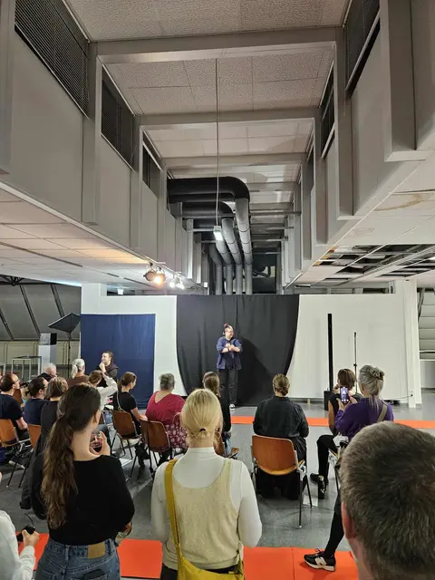 Kurator*in Oliver Suchanek bei der Eröffnungsfeier

Oliver steht vor einer schwarzen Wand, davor sieht man einige Personen vom Publikum von hinten.