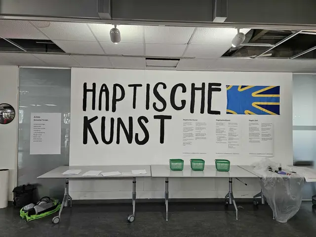 Eine Wand mit der Aufschrift "Haptische Kunst", daneben hängt eine Deaf Flag (eine Flagge mit einer hellblauen/türkisen Hand mit gelber Umrandung auf dunkelblauem Hintergrund. 
Daneben hängt Information zur Ausstellung auf deutsch, leichter Sprache und englisch. Es ist zu klein, um es auf dem Foto lesen zu können.
Davor stehen mehrere graue Tische, auf denen Papier liegt und drei leere grüne Körbe. 