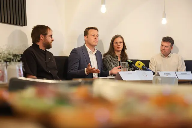 Vier Personen, drei Männer und eine Frau, sitzen an einem Tisch. Vor ihnen sind zwei Mikros aufgebaut, es handelt sich um eine Pressekonferenz.