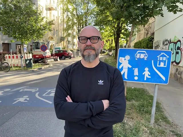 Der KPÖ-Politiker Hanno Wisiak steht vor einem Wohnstraßenschild. 