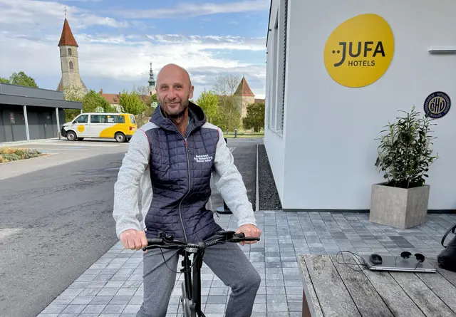 Ein Mann im mittleren Alter auf dem Fahrrad sitzend blickt freundlich in die Kamera, im Hintergrund der Eingang zum JUFA Hotel.