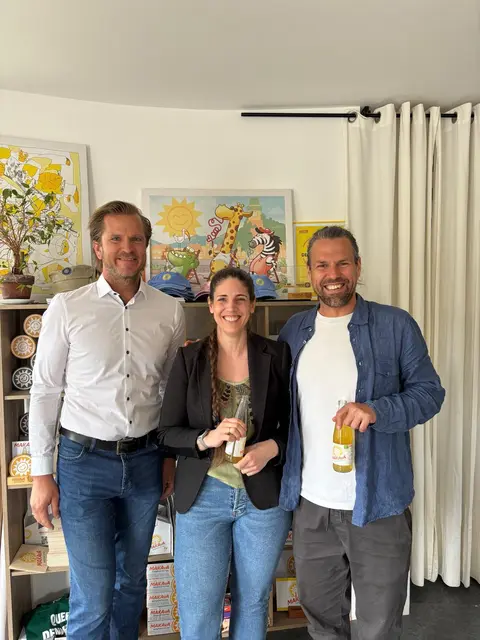 Zwei Männer an den Seiten und in der Mitte eine Frau. Die zwei Personen links halten eine Flasche Makava in der Hand