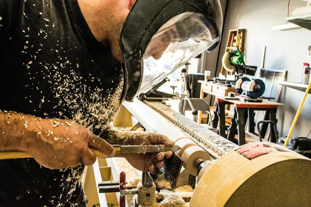 Ein Tischler arbeitet in einer Werkstatt an einem Holzstück, während Holzspäne durch die Luft fliegen. Die Szene vermittelt handwerkliche Präzision, Kreativität und die Arbeit an Projekten für die Tischler Trophy 2025/26.