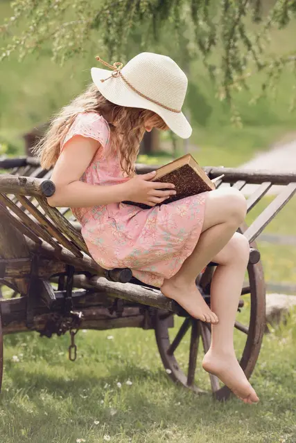 Ein Mädchen sitzt in einem Kleid auf einem landwirtschaftlichen Gerät und liest ein Buch.