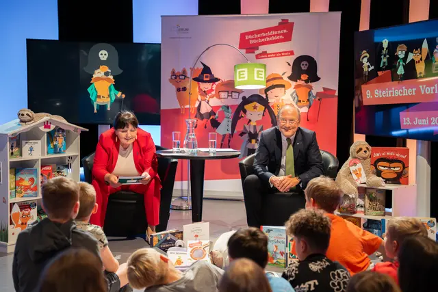 Eine Frau und ein Mann sitzen auf Stühlen vor einer Gruppe Schulkindern.