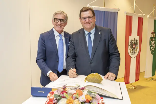 Zwei Männer, einer davon mit einem Stift in der Hand, stehen vor einem großen Buch und lächeln in Richtung Kamera. 