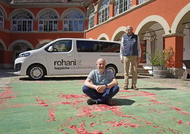 Zwei Männer, einer sitzend und einer stehend auf einem Riesenteppich im Innenhof eines Schlosses, lächeln freundlich in die Kamera, während im Hindergrund ein Bus mit dem Firmenlogo Rohani steht.