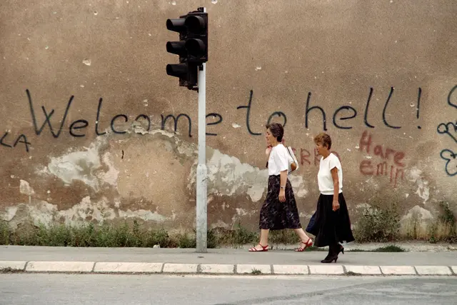 Zwei Frauen gehen am 24. August 1993 durch den als „Sniper Alley“ bekannten Stadtteil von Sarajevo, in der Nähe einer Wand mit der Graffitibeschriftung „Willkommen in der Hölle“. | Foto: APA-Images / AFP / GABRIEL BOUYS
