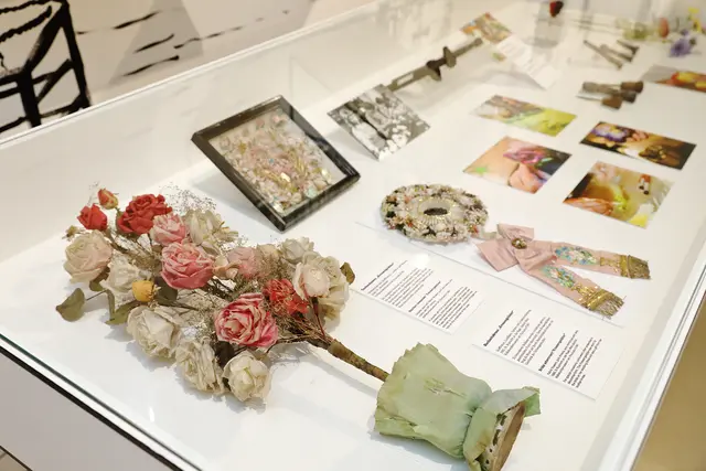 Austellungsstücke in einem Glaskasten: Ein Strauß aus künstlichen Blumen, Schmuck und Fotos. DIe Fotos zeigen die Herstellung von Kunstblumen.