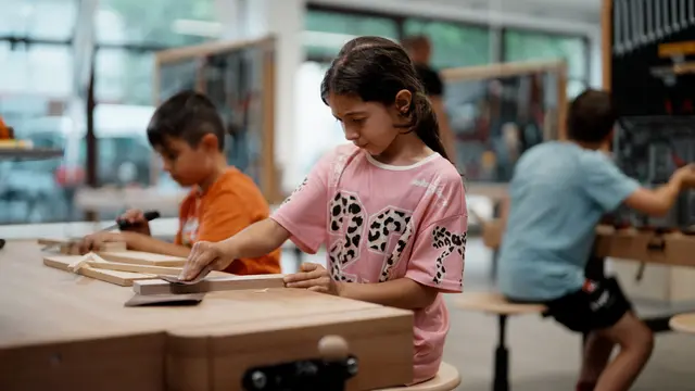 Kinder sitzen an Werkbänken und bearbeiten Holzstücke mit verschiedenen Werkzeugen.