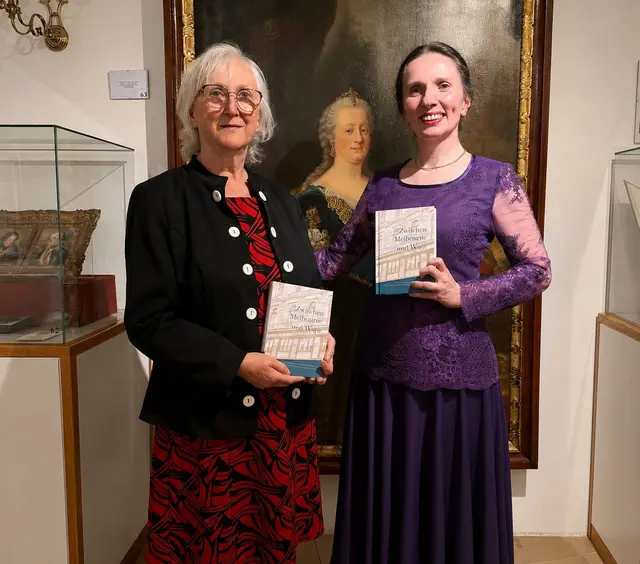 Verlegerin Margarete Tischler (links) und Autorin Johanna Beisteiner (rechts) stehen nebeneinander und blicken in die Kamera. Im HIntergrund ist ein Gemälde der Kaiserin Maria Theresia zu sehen. Urheber: Kulturverein K.U.N.