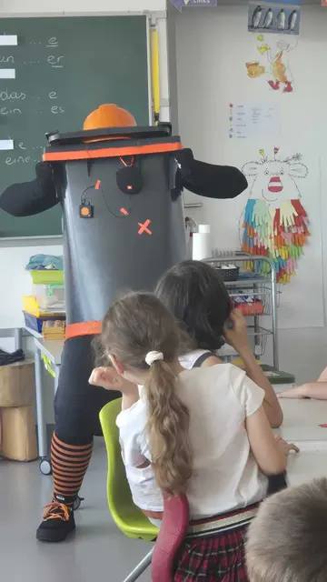 Die Schauspielerin versteckt sich im Mülltonnenkostüm, indem sie den Kopf einsteckt; davor ein paar Kinder aus der Schulklasse.