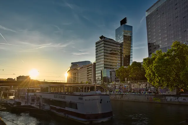 Blick auf den Wiener Donaukanal bei Sonnenuntergang mit dem Fahrgastschiff „Blue Danube“ im Wasser. Links steht die echte Sonne tief am Himmel und erzeugt Gegenlicht und Blendenflecke. Rechts spiegeln sich die Sonnenstrahlen gleißend hell in einem Wolkenkratzer mit Glasfront. Am gegenüberliegenden Ufer sind begrünte Bäume und Mauern mit Graffitis zu sehen.