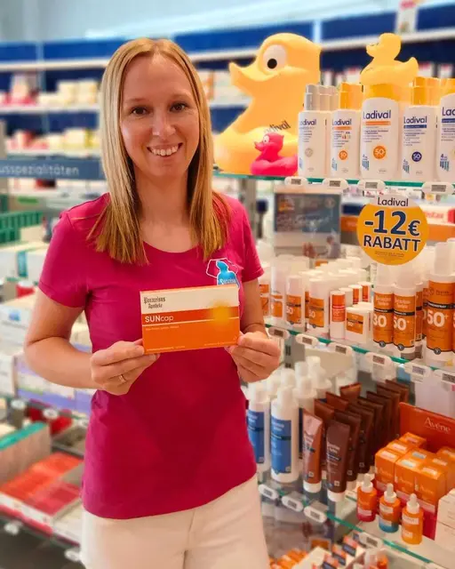 Julia von der Paracelsus-Apotheke Weiz steht vor Sonnenpflegeprodukten und hält eine SUNcap-Packung.