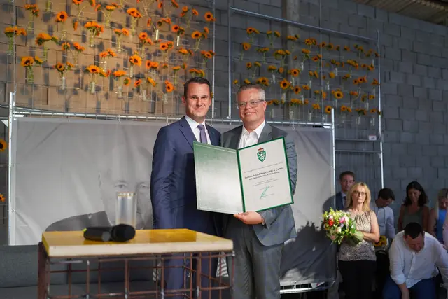 Feierliche Übergabe einer Auszeichnung an eine Person. Im Hintergrund eine Wand mit floraler Dekoration.