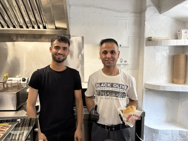 2 Männer stehen in einer Küche, einer hält einen Kebap