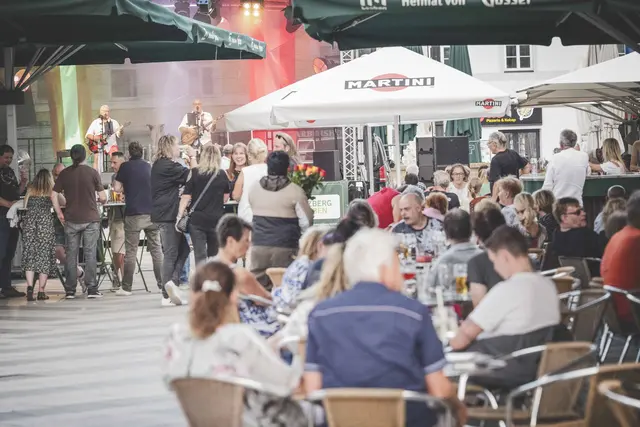 Viele Menschen stehen vor einer Bühne und sitzen im Gastgarten