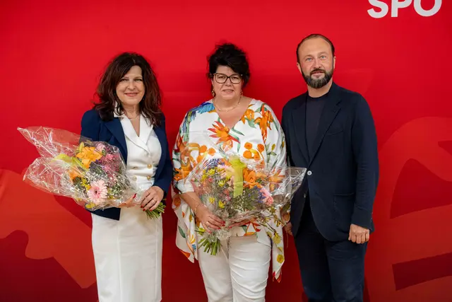 2 Frauen und 1 Mann stehen vor einem roten Transparent. Die 2 Frauen halten Blumen