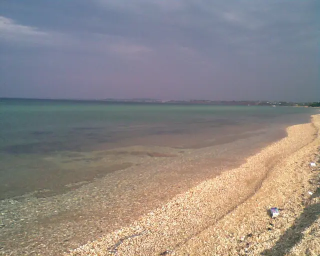 Grober Sandstrand und dunkelgrünes Meer. Im Hintergrund Land. 