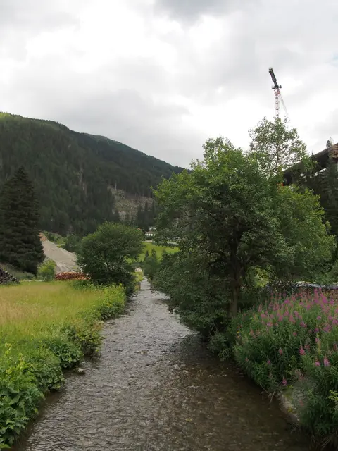 Ein kleiner, flacher Fluss fließt durch ein Tal mit grünen Wiesen, im Hintergrund ist ein gelber Baukran sichtbar.