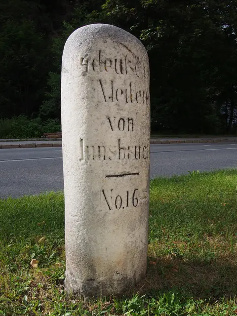 Historischer Meilenstein aus Stein mit der Inschrift 4 deutsche Meilen von Innsbruck am Straßenrand vor unscharfem Hintergrund.