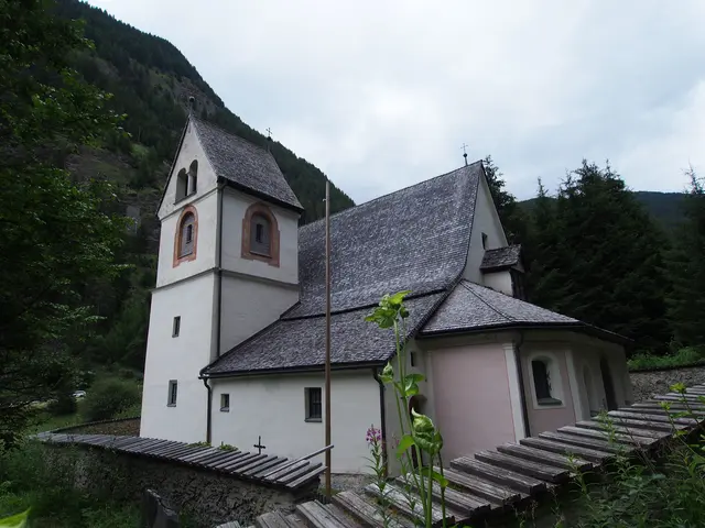 Detailaufnahme einer weißen Kirche mit Holzschindeldach und rosa Verzierungen vor bewaldeten Berghängen.