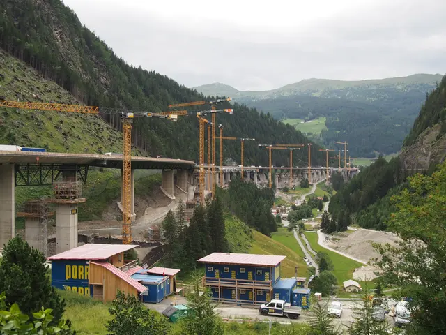 Panorama einer Großbaustelle im Tal mit einer Reihe gelber Baukräne, Betonpfeilern und blauen Baustellenelementen im Wald.