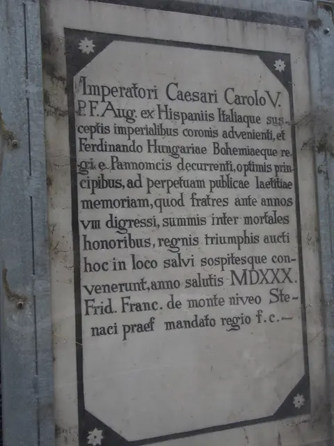 In eine graue Felswand gemeißelte Gedenktafel mit lateinischer Inschrift hinter einer transparenten Schutzabdeckung.